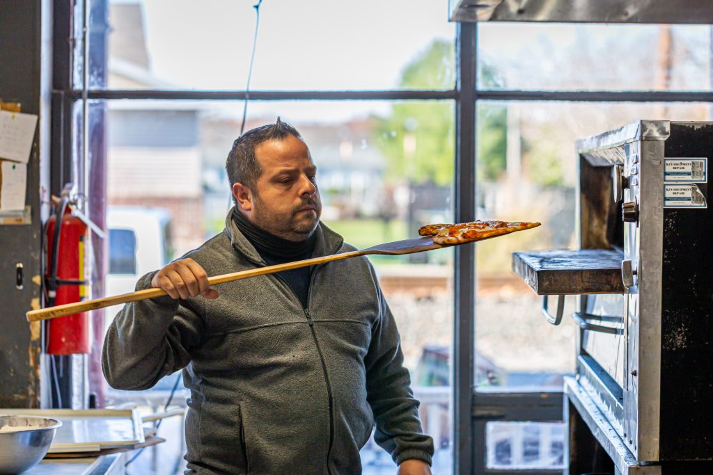 A man holding a wooden spatula in front of a pizza oven.